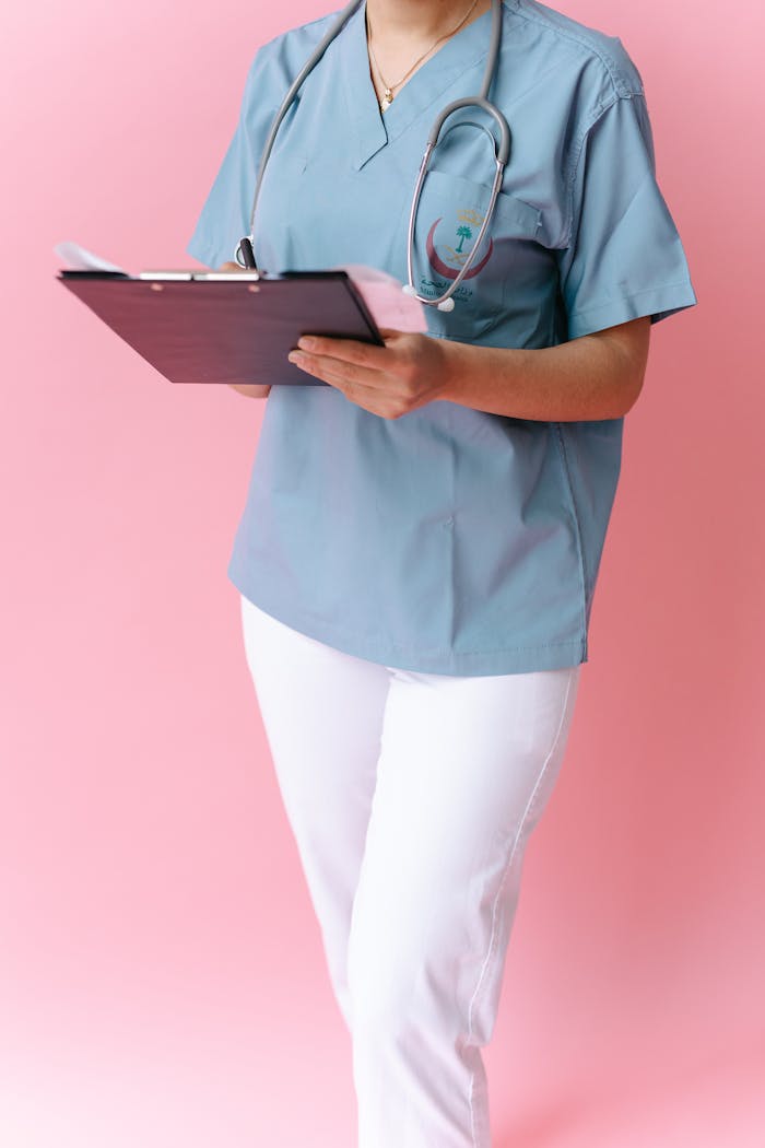 Nurse in blue scrubs with a stethoscope holding a clipboard against a pink backdrop.
